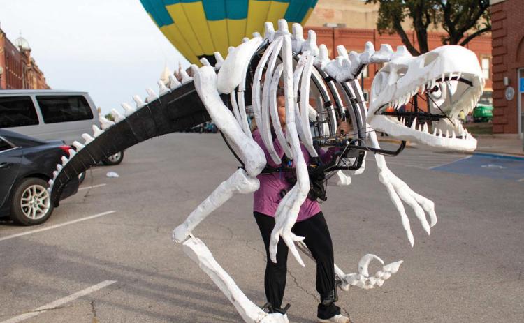 A towering dinosaur roams downtown Guthrie on Halloween, with a hot air balloon providing a dramatic backdrop for trick-or-treaters.